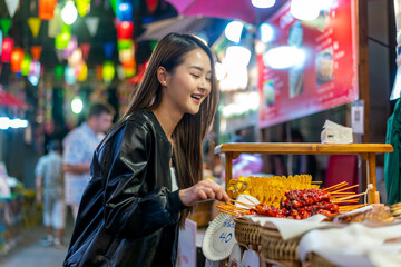 Asian woman enjoying street food at Chiang Mai Walking Street Night Bazaar. Young traveler exploring vibrant night market in Thailand.