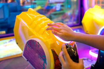 Close-up of a child's hand touching a glowing button on a bright yellow dinosaur-themed arcade machine, surrounded by colorful lights and gaming screens in an indoor play center.
