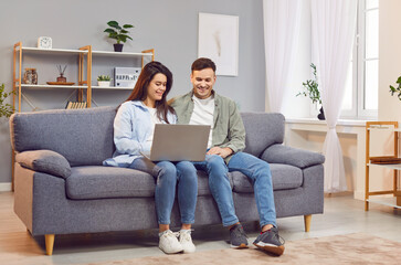 Happy couple at home, young woman and man sitting close in new modern living room, have fun using...