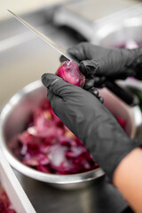 Hands in black gloves peeling a red onion with a knife over a metal bowl, showcasing hygienic food...