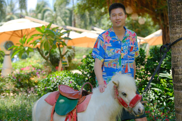 Asian man in a floral shirt petting a white pony miniature horse