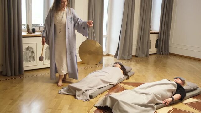 A meditation guide conducts a gong meditation session with two participants lying on mats. The serene atmosphere and focused energy create a peaceful environment for deep relaxation.