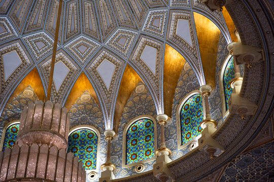 An ornate ceiling with stained glass windows, intricate patterns, and a large chandelier. No people are present