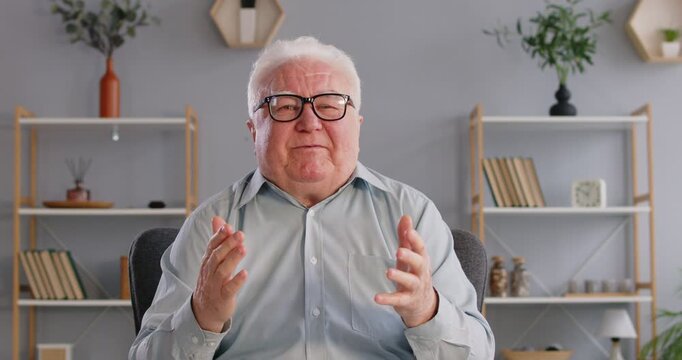 Elderly man sits in front of camera and addresses audience with important message of life experience. Webcam view of friendly Caucasian gray-haired old man in shirt and glasses telling stories.