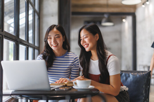 Two young students smiling and studying together with laptop and book in cafe