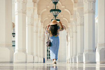 A woman walking under stone arches, wearing a floral blouse, blue skirt, and wide-brimmed hat,...