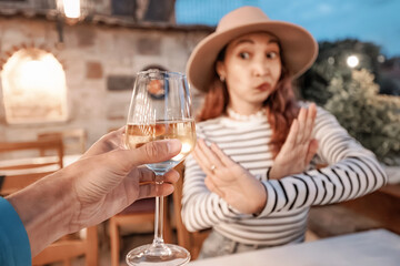 Woman with hat refusing alcohol making stop gesture with hands at restaurant table, with glass of white wine in the foreground