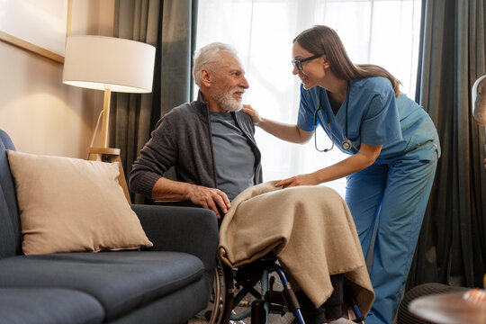 Smiling, beautiful nurse covering elderly man in wheelchair with blanket at home