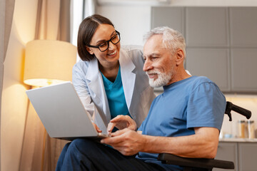 Doctor assisting senior man in wheelchair using laptop at home