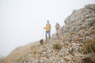 Two Travelers Explore Rocky Terrain with a Dog Amid a Misty Landscape