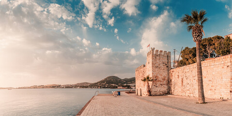 Foca castle stands majestically by the calm sea under a vibrant sky, with palm trees and mountains...