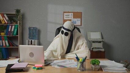 Halloween businessman ghost in white sheet and necktie over head sits at office desk works on...
