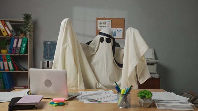 Halloween businessman ghost in white sheet with necktie over head sits at office desk having party dancing in chair. Happy halloween business concept.