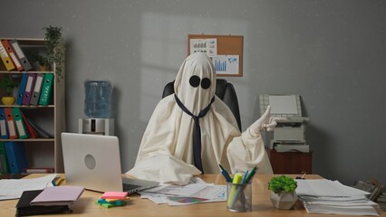 Halloween businessman ghost in white sheet sits at office desk looks at camera holding hand up and pointing finger at side. Happy halloween business concept.