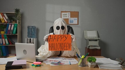 Halloween businessman ghost in white sheet sits at office desk holds shows orange paper sign happy...