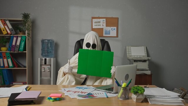 Halloween businessman ghost in white sheet sits at office desk holding showing chroma key green screen board at camera. Happy halloween business concept.