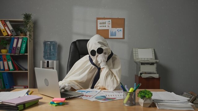 Halloween businessman ghost in white sheet sits at office desk works on laptop, tired bored expression, sighing and thinking. Happy halloween business concept. - Powered by Adobe