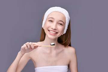 Smiling caucasian young female holding bamboo toothbrush with charcoal paste against gray background.