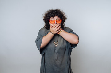 Person with curly hair and orange glasses covers mouth in playful surprise against a plain...