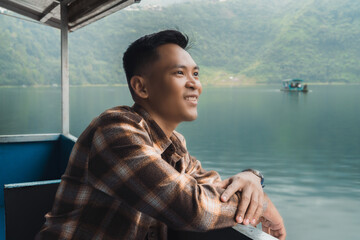 Asian young man on a boat overlooking a serene lake surrounded by lush green hills and a misty atmosphere, epitomizing joy, relaxation, and a connection with nature.
