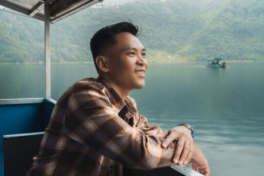 Asian young man on a boat overlooking a serene lake surrounded by lush green hills and a misty atmosphere, epitomizing joy, relaxation, and a connection with nature.
