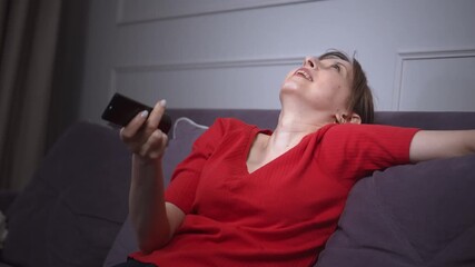 Young woman in red shirt sitting on sofa holding remote control and watching TV, her facial expression changing while navigating the channels