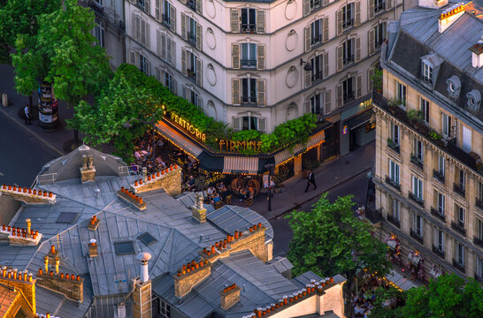 Aerial view of a bustling Parisian cafe with illuminated signs and rooftops with typical French chimneys at dusk.