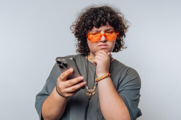 Young person with curly hair and orange glasses contemplating a message on their smartphone indoors