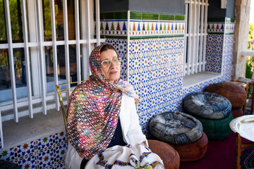 Elderly Arab woman with hijab sits calmly on a porch. Surrounding her are decorative Moroccan tile...