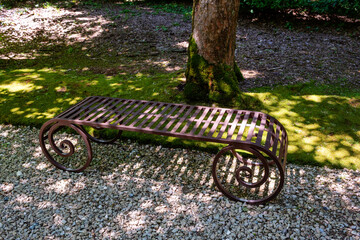 Ornate metal bench featuring scrollwork design is situated on gravel, surrounded by vibrant greenery and trees, providing a tranquil outdoor atmosphere. 