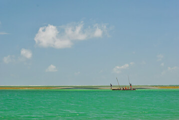 Obraz premium A group of people standing on a traditional wooden boat in turquoise water near a sandy shore, under a clear blue sky with scattered clouds.