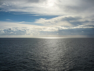 Vast open sea under dramatic clouds with sunlight breaking through, creating a silver reflection on the ocean surface