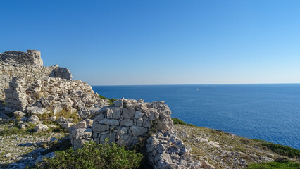 Stone ruins on a rocky hill overlooking the Adriatic Sea with sailboats in the distance, under a clear blue sky in Kornati National Park, Croatia.