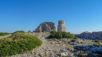 Fototapeta premium Ruins of an ancient stone church with a preserved arch and bell tower on a rocky hill in Kornati National Park, Croatia, under a bright blue sky.