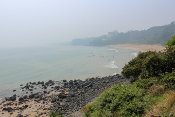 View of Jungmun Beach coastline on Jeju Island, South Korea, with surfers in the water, rocky shore, hotel buildings in the distance, and misty atmosphere
.