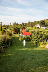 A child in light clothing and a sun hat walks on a grassy path bordered by flowers. Red roofed...