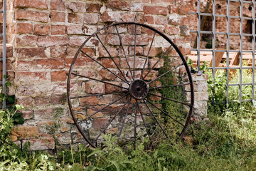 Old rusty metal wagon wheel resting against weathered brick wall, surrounded by overgrown grass and...