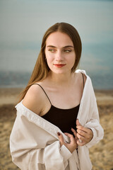 Young woman stands by serene beach, draped in loose white shirt. Her gaze reflects contemplation...