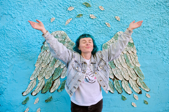 Fototapeta Young woman with dyed hair poses joyfully in front of blue mural. Angel wings painted on wall create illusion of wings on her back. Denim jacket enhances casual look