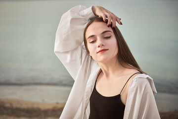 Young woman in white shirt and black top poses with confidence by tranquil seaside. Her relaxed...