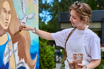 Young adult female in casual clothing actively paints large mural outdoors. She holds brush near...