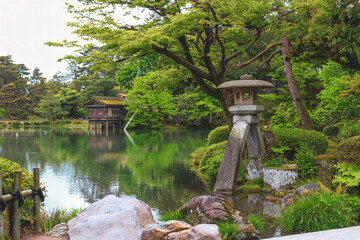 Traditional Japanese stone lantern by a pond in Kenrokuen Garden, Kanazawa, with lush greenery and a wooden teahouse in the background.