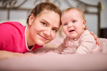 Young mother with brown hair and baby girl lying on pink blanket. Warm lighting, indoor setting,...