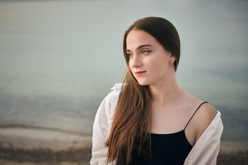 Young woman stands contemplatively by serene seaside, with gentle waves in background. Her relaxed...