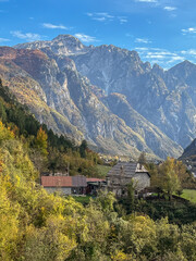 Traditional rural house in northern Albania surrounded by colorful autumn forest, with dramatic mountains in the background under a partly cloudy sky.