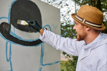 Young male artist in straw hat, white coat painting outdoors. Canvas features abstract figure with...
