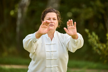 Mature woman, brown hair, caucasian, focuses on qigong. She wears traditional white attire. Sunlit...