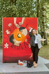 Young woman with light brown hair and casual clothes stands by large surreal painting in park....