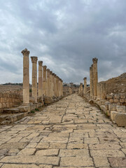 Ancient Roman colonnaded street in Jerash, Jordan, with weathered stone pavement and cloudy sky,...