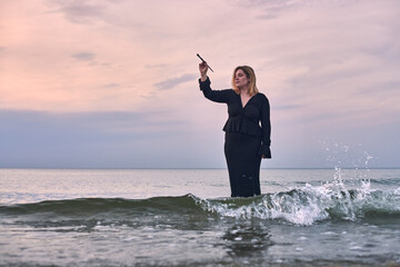 Young woman stands waist-deep in ocean during sunset, holding paintbrush. Serene seascape evokes...